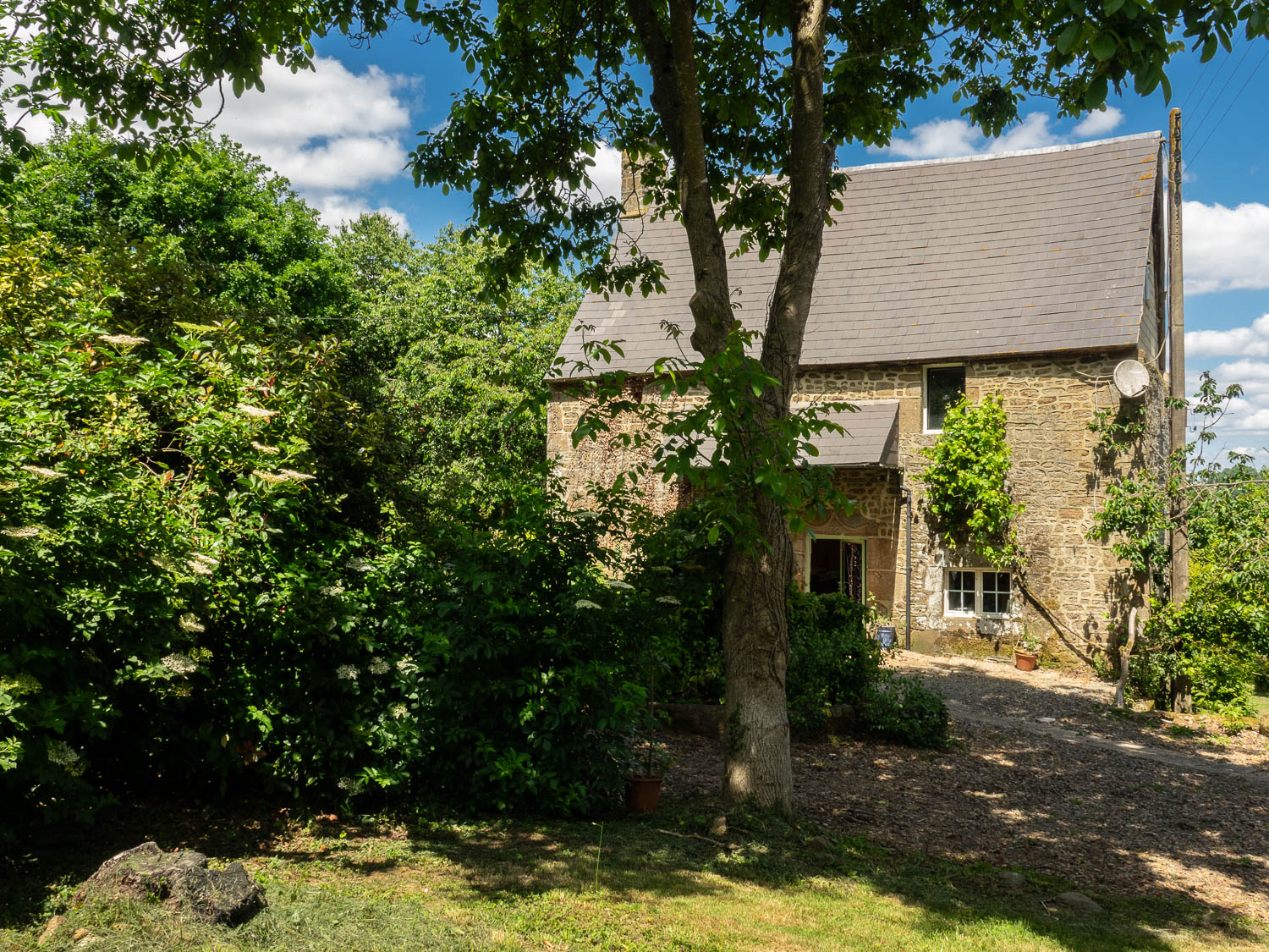 Photograph of an old stone-built house in the sunshine under blue skies and surrounded by trees and shrubs.